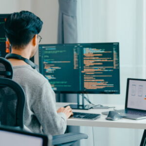 Software developer working at a desk with dual monitors displaying code and a laptop showing a user interface design.