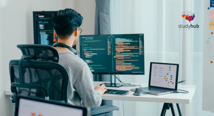 Software developer working at a desk with dual monitors displaying code and a laptop showing a user interface design.