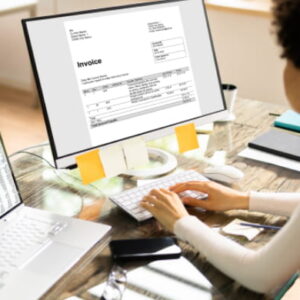 Professional working at a modern desk reviewing an invoice on a desktop monitor with a spreadsheet open on a laptop.