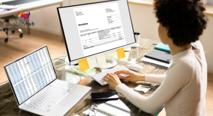 Professional working at a modern desk reviewing an invoice on a desktop monitor with a spreadsheet open on a laptop.