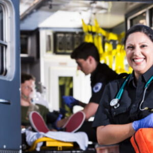 A smiling female paramedic in uniform holding a medical bag in front of an open ambulance with a colleague treating a patient in the background.
