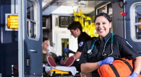 A smiling female paramedic in uniform holding a medical bag in front of an open ambulance with a colleague treating a patient in the background.