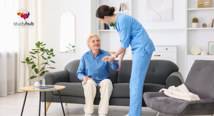 Female nurse in blue scrubs assisting an elderly woman to stand up from a sofa in a home care setting.
