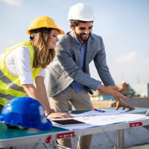 Two construction professionals wearing safety helmets reviewing building plans and a laptop at an outdoor construction site.