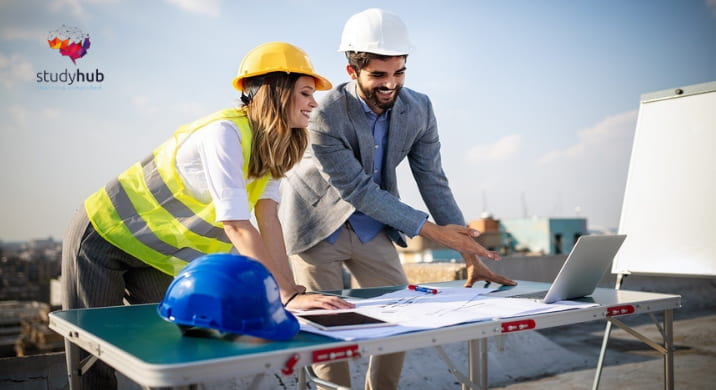 Two construction professionals wearing safety helmets reviewing building plans and a laptop at an outdoor construction site.