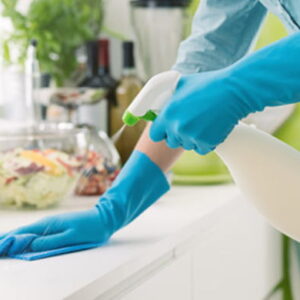 Person wearing blue gloves cleaning a kitchen countertop with a spray bottle and microfiber cloth.
