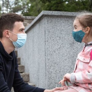 Adult wearing a face mask talking with a young schoolgirl wearing a backpack and mask outdoors on stone steps.