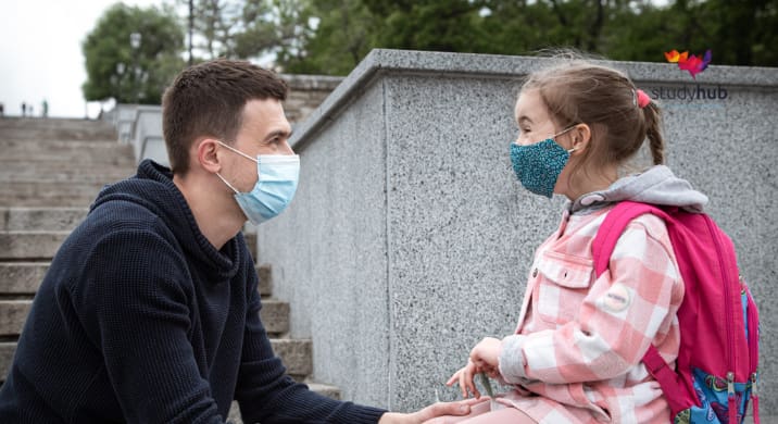Adult wearing a face mask talking with a young schoolgirl wearing a backpack and mask outdoors on stone steps.
