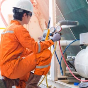 Industrial technician in orange protective coveralls inspecting pressure gauges on mechanical equipment.