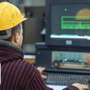 Industrial worker wearing a yellow safety helmet operating a CNC machine and monitoring a computer screen in a manufacturing workshop.