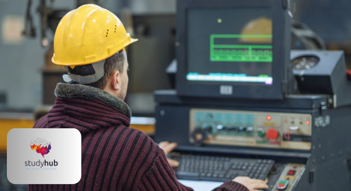 Industrial worker wearing a yellow safety helmet operating a CNC machine and monitoring a computer screen in a manufacturing workshop.