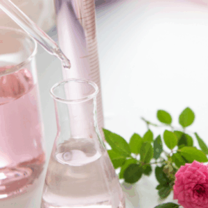 Close-up of a glass pipette dripping liquid into a flask containing pink fragrance, sitting next to a fresh pink rose.