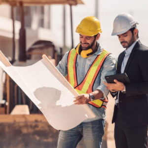 Construction engineers reviewing building plans at an active construction site while wearing safety helmets.