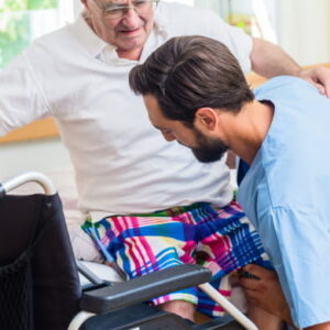 Healthcare professional assisting elderly man from wheelchair in hospital room