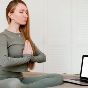 Young woman practicing meditation in a seated yoga pose with eyes closed, using a laptop for online yoga or mindfulness training at home.