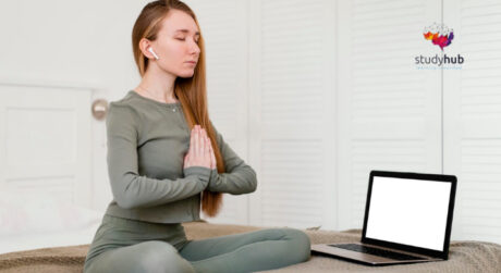 Young woman practicing meditation in a seated yoga pose with eyes closed, using a laptop for online yoga or mindfulness training at home.