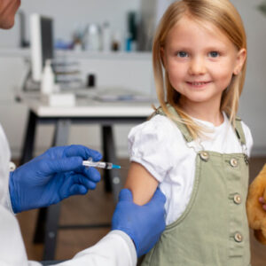 Young child holding a teddy bear while receiving a vaccination from a healthcare professional.