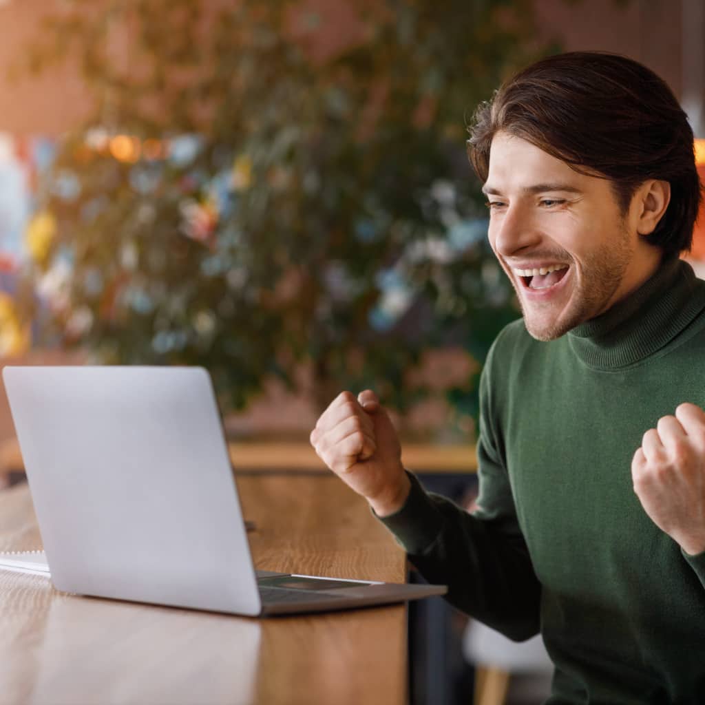 Happy man celebrating success while working on a laptop at a desk in a modern workspace.
