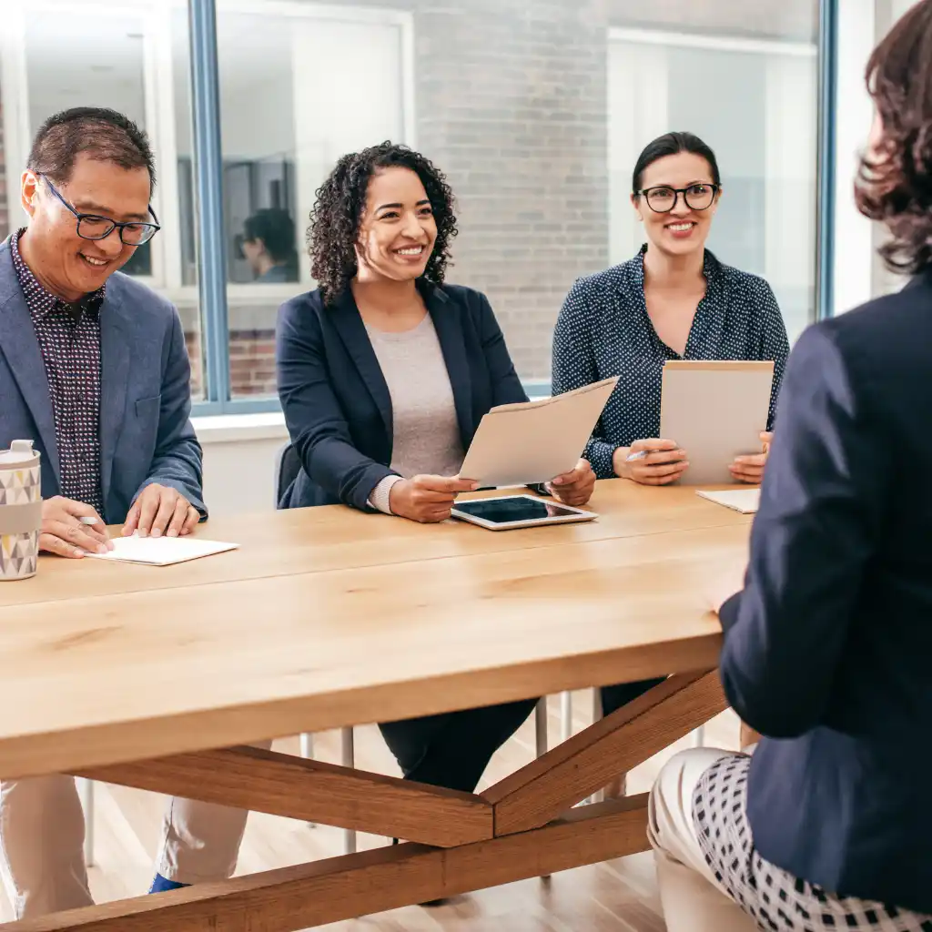 A diverse group of professionals sitting at a table during a job interview, reviewing documents and smiling at a candidate in a modern office.