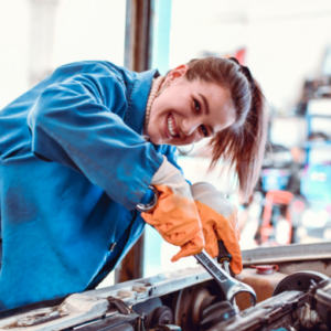 Smiling female mechanic in blue uniform using a wrench to repair a car engine in an auto workshop