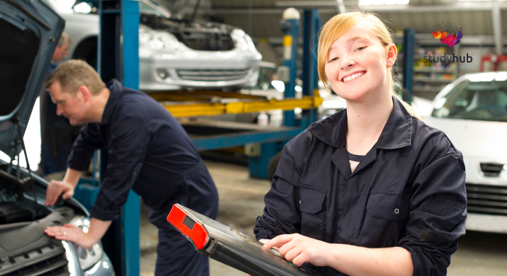 Smiling female auto mechanic holding a clipboard in a car repair workshop while a colleague works under the hood of a vehicle in the background.