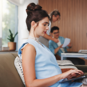 Pregnant woman working on a laptop from home while colleagues collaborate in the background