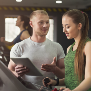Personal trainer guiding a young woman on a treadmill while holding a tablet in a modern gym.
