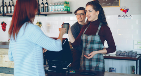 Barista serving takeaway coffee to a customer at a café counter while another staff member stands in the background.