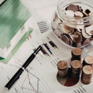 Coins stacked beside a glass jar, calculator, and model house on financial charts and reports