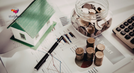 Coins stacked beside a glass jar, calculator, and model house on financial charts and reports