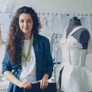 Fashion designer standing in studio with dress form and measuring tape, surrounded by sketches and sewing equipment