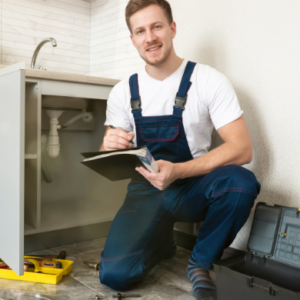 Plumber kneeling by an open kitchen cabinet inspecting pipes and taking notes with tools nearby
