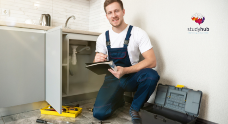Plumber kneeling by an open kitchen cabinet inspecting pipes and taking notes with tools nearby
