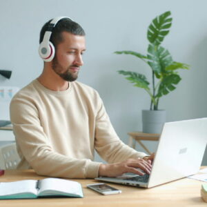 Man wearing headphones working on a laptop at a tidy home office desk with notebook, phone, and plants.